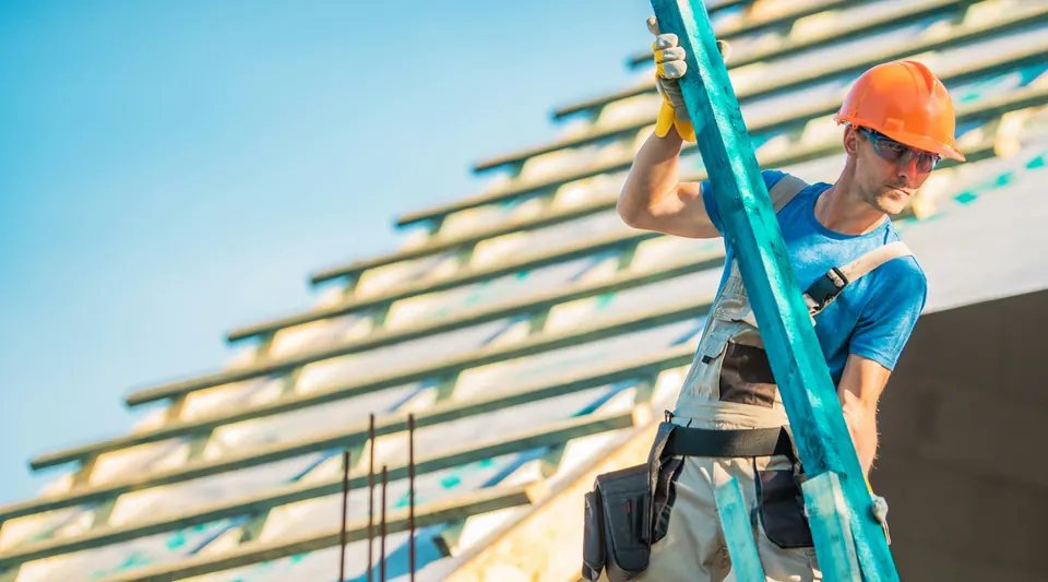 Roofer in a hard hat carrying lumber across an open roof frame